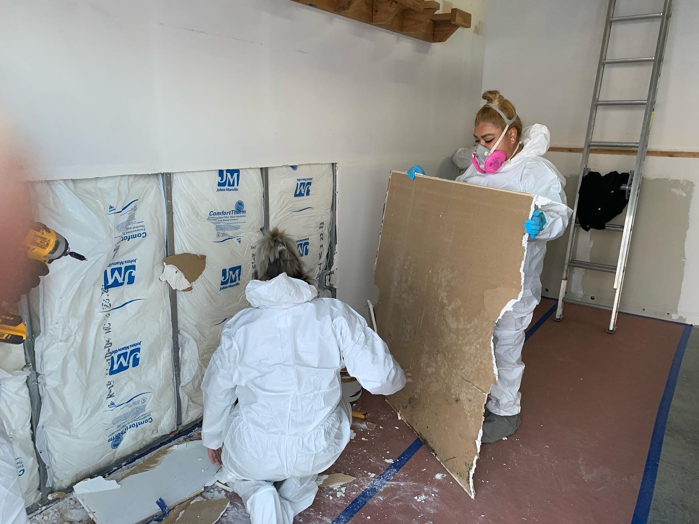 Two people in protective suits and masks removing old insulation from a wall in a room under renovation.