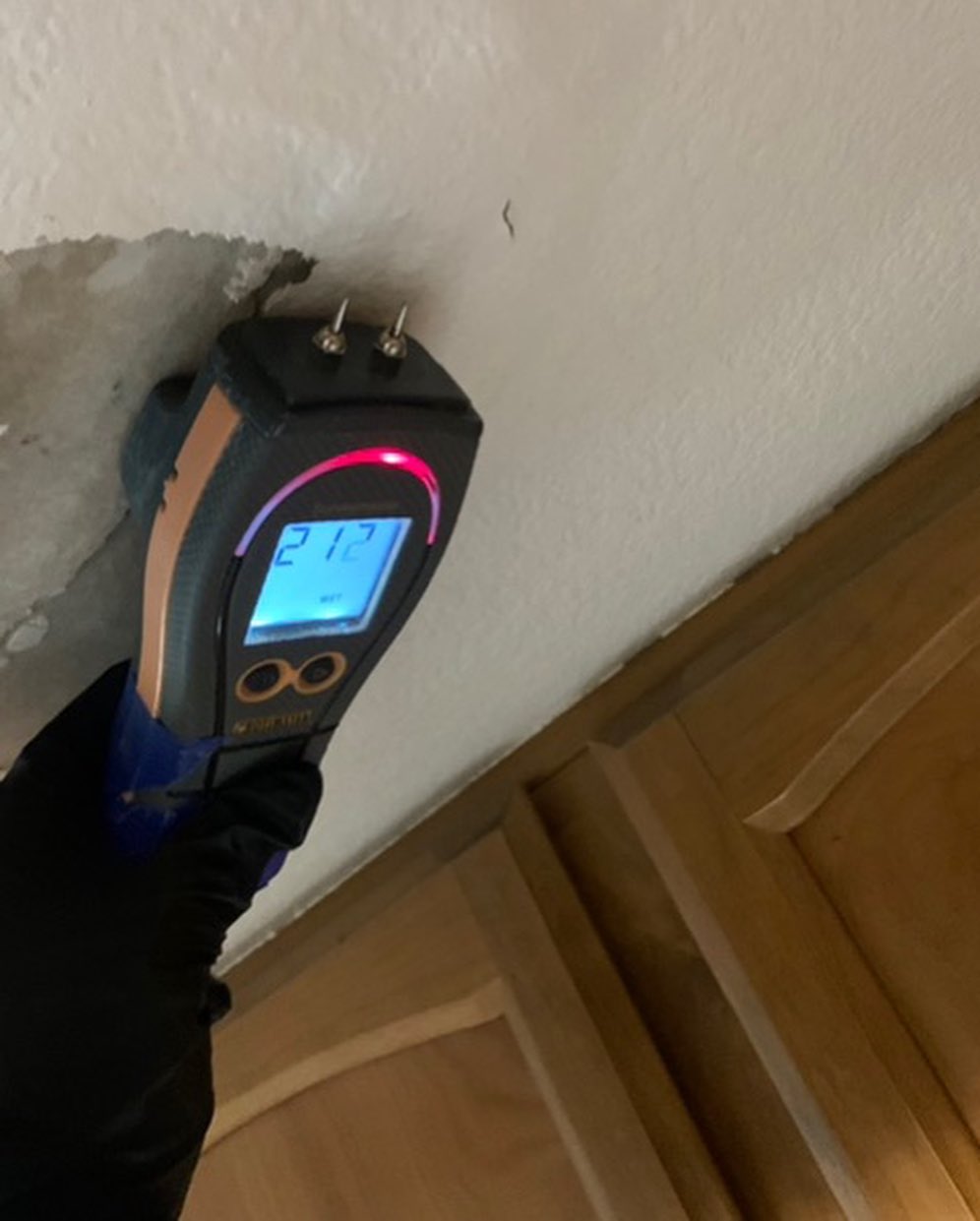 A hand holding a digital moisture meter against a damaged, peeling wall, displaying a reading of 21.2%.