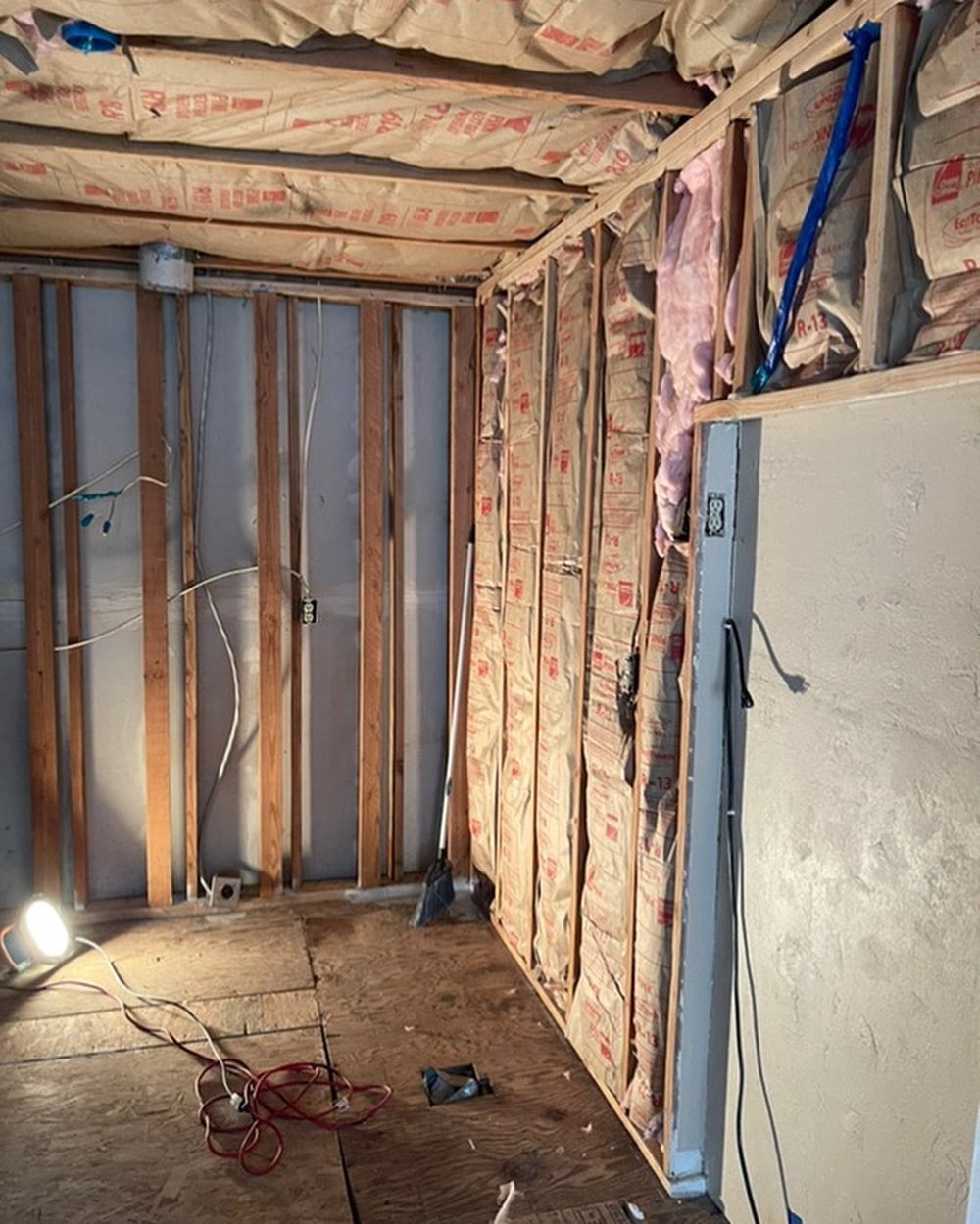 A partially renovated room with exposed wall studs and insulation, electrical wiring, a work light, and a broom shows signs of water damage restoration. Some areas of the wall remain unfinished.