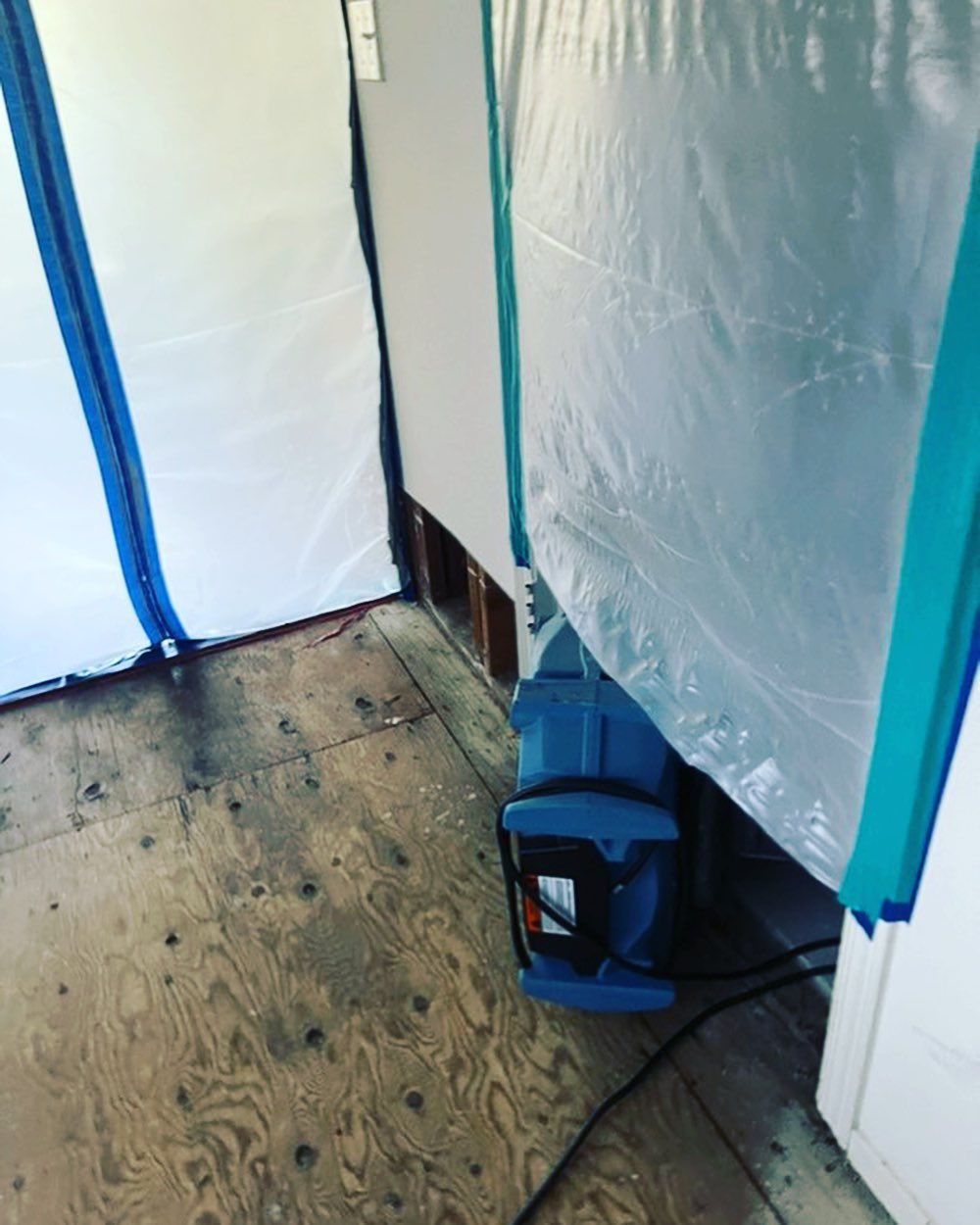A room undergoing renovation with protective plastic sheeting covering the walls and floor suggests active mold restoration. A small section of drywall has been removed, revealing exposed studs, and a piece of equipment is on the floor.