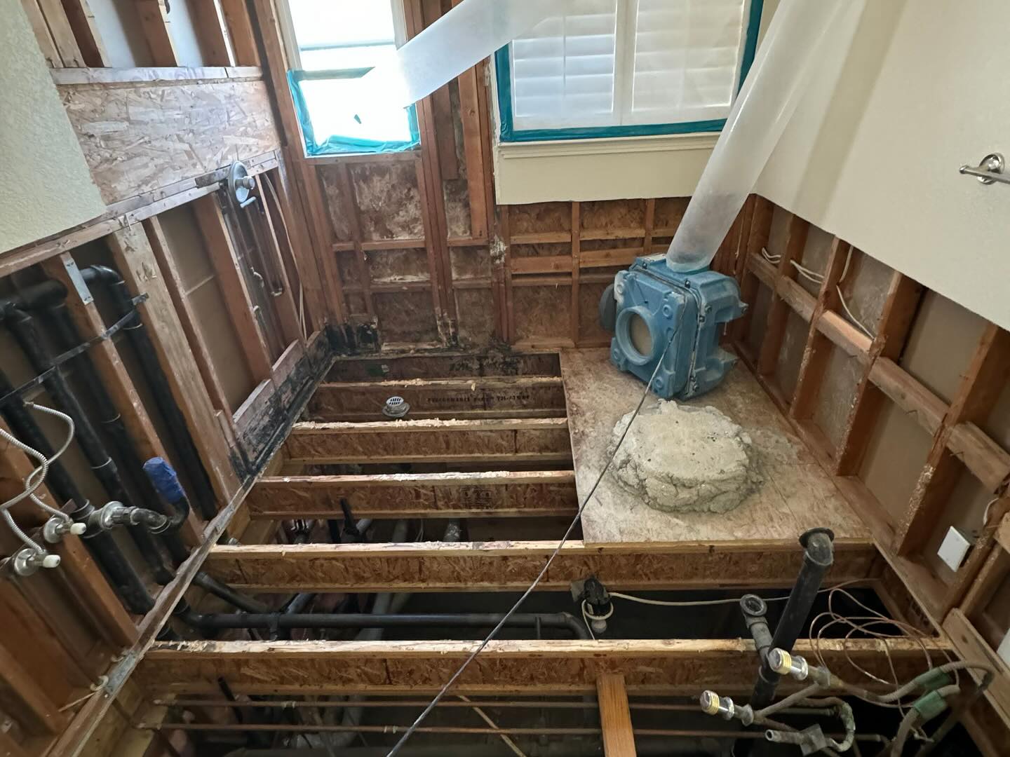 Interior of a building under renovation, showing exposed wooden beams, plumbing pipes, and a dismantled floor with a visible concrete foundation.