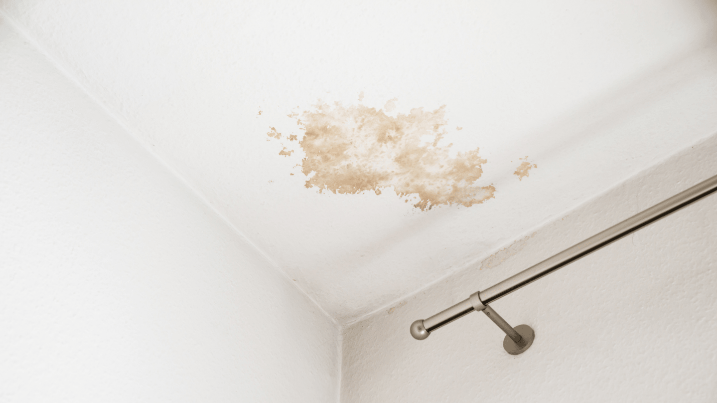 A white ceiling with a large brown water stain near the corner, a clear sign of water damage, adjacent to a metal curtain rod attached to the wall.