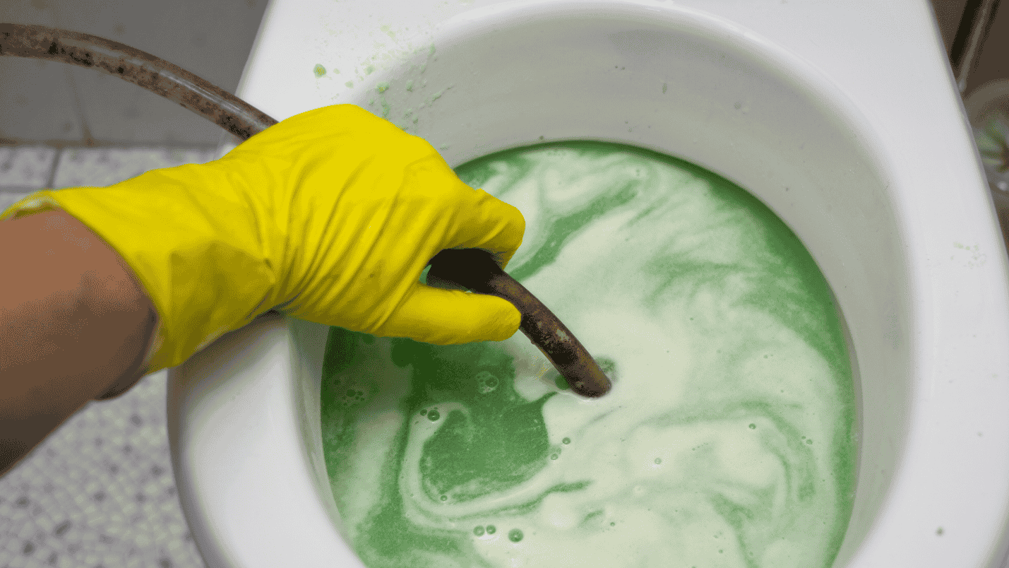 A gloved hand holds a tube down a toilet that is full of green liquid.