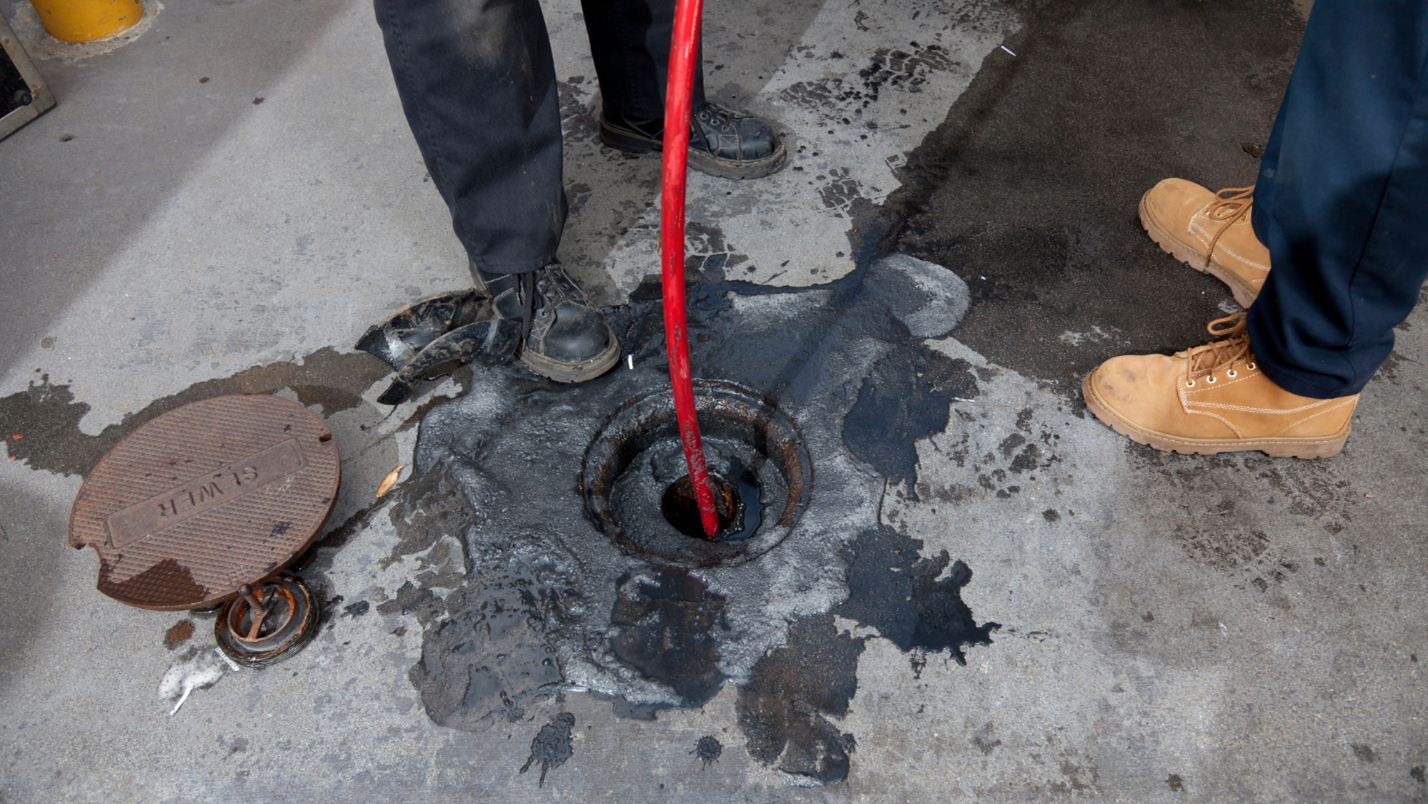 Two workers stand next to a sewer drain and send a long tool into the drain.